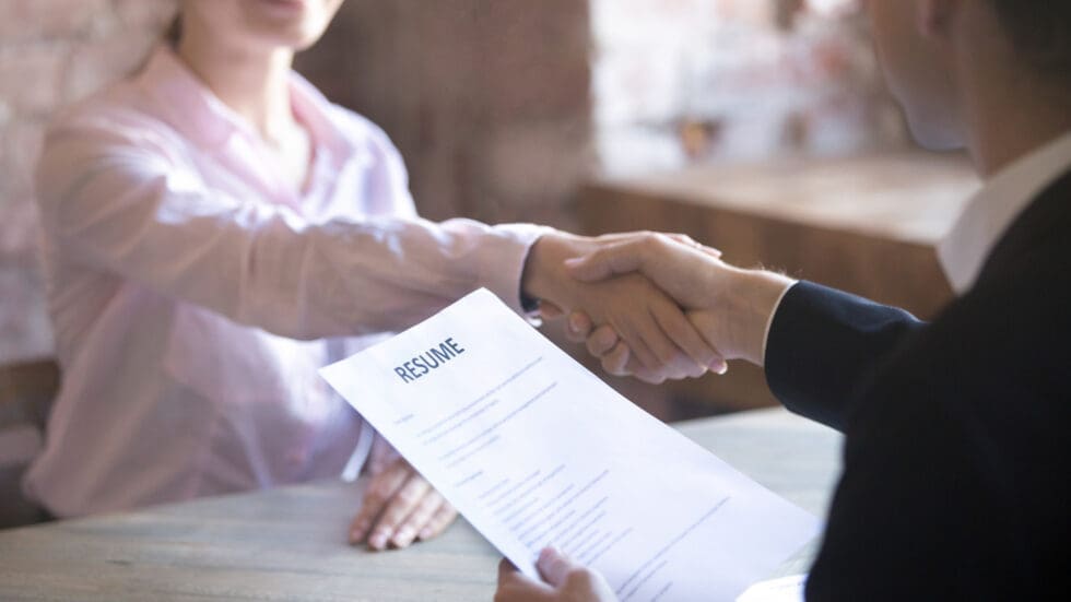 A job candidate and an interviewer shake hands across a table while one holds a printed resume, in a warmly lit office or café setting.