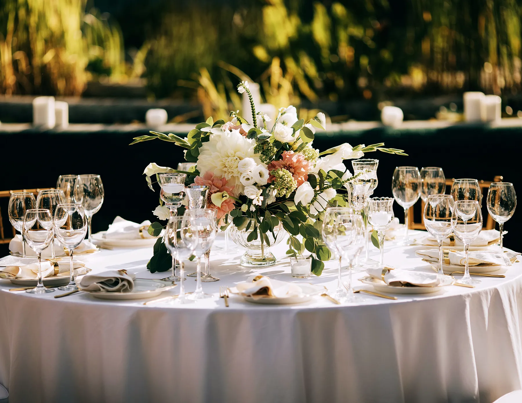 An elegantly set outdoor wedding reception table with a white linen, crystal glassware, gold cutlery, and a lush centerpiece of peach and white blooms with eucalyptus greenery.