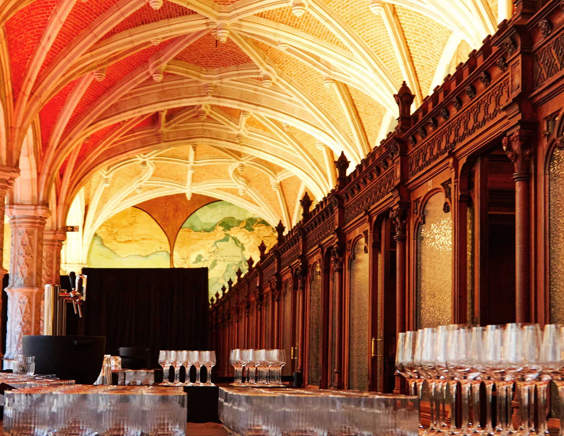 Rows of crystal glassware and beer taps line a bar setup inside a magnificent Gothic-vaulted hall with ornate dark wood paneling and a large world map mural illuminated at the far end.