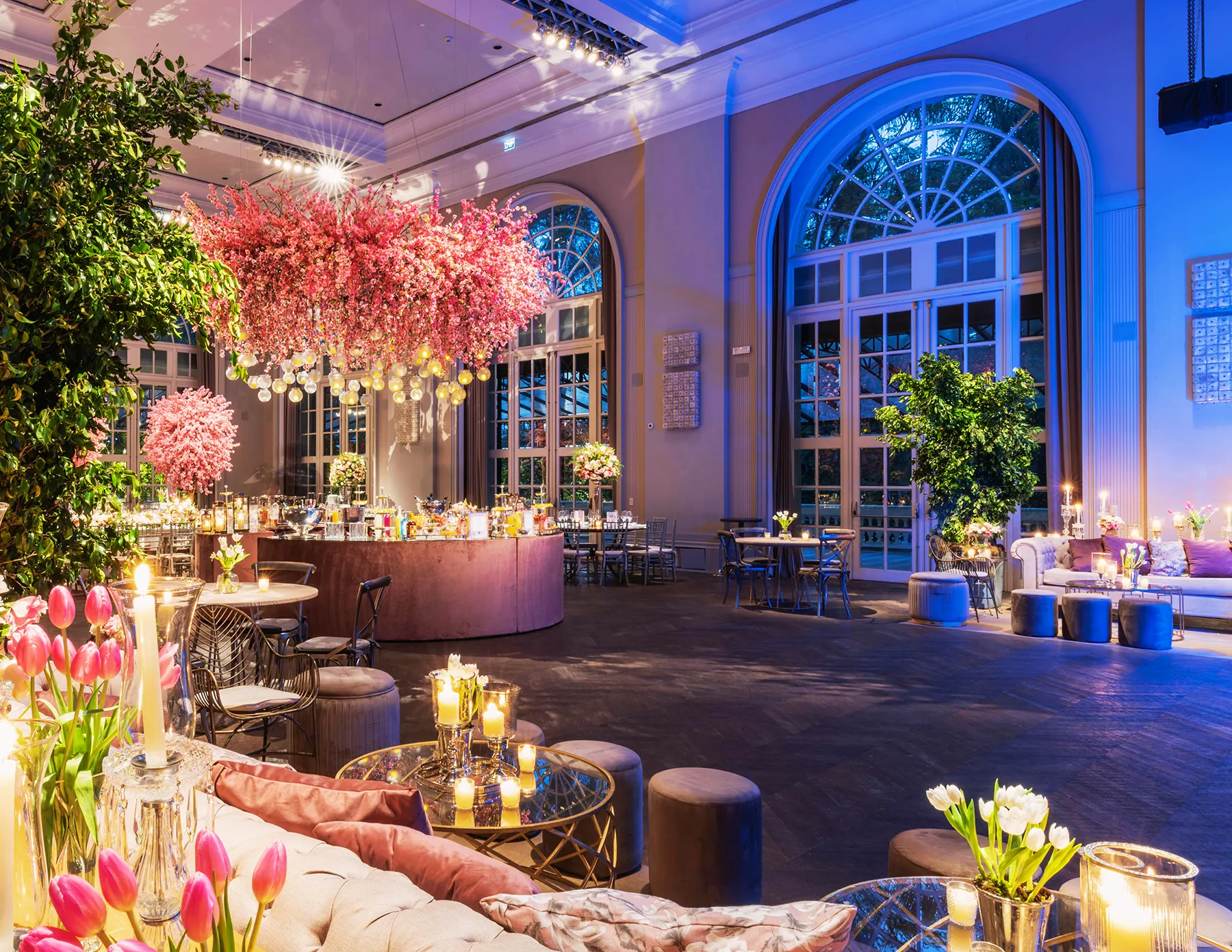 elegant-garden-party-venue-cherry-blossom-blue-lighting-hmg-plus. A luxurious event venue interior featuring a dramatic suspended cherry blossom installation, a curved bar, candlelit lounge seating, pink tulips, and tall arched windows bathed in blue evening light.