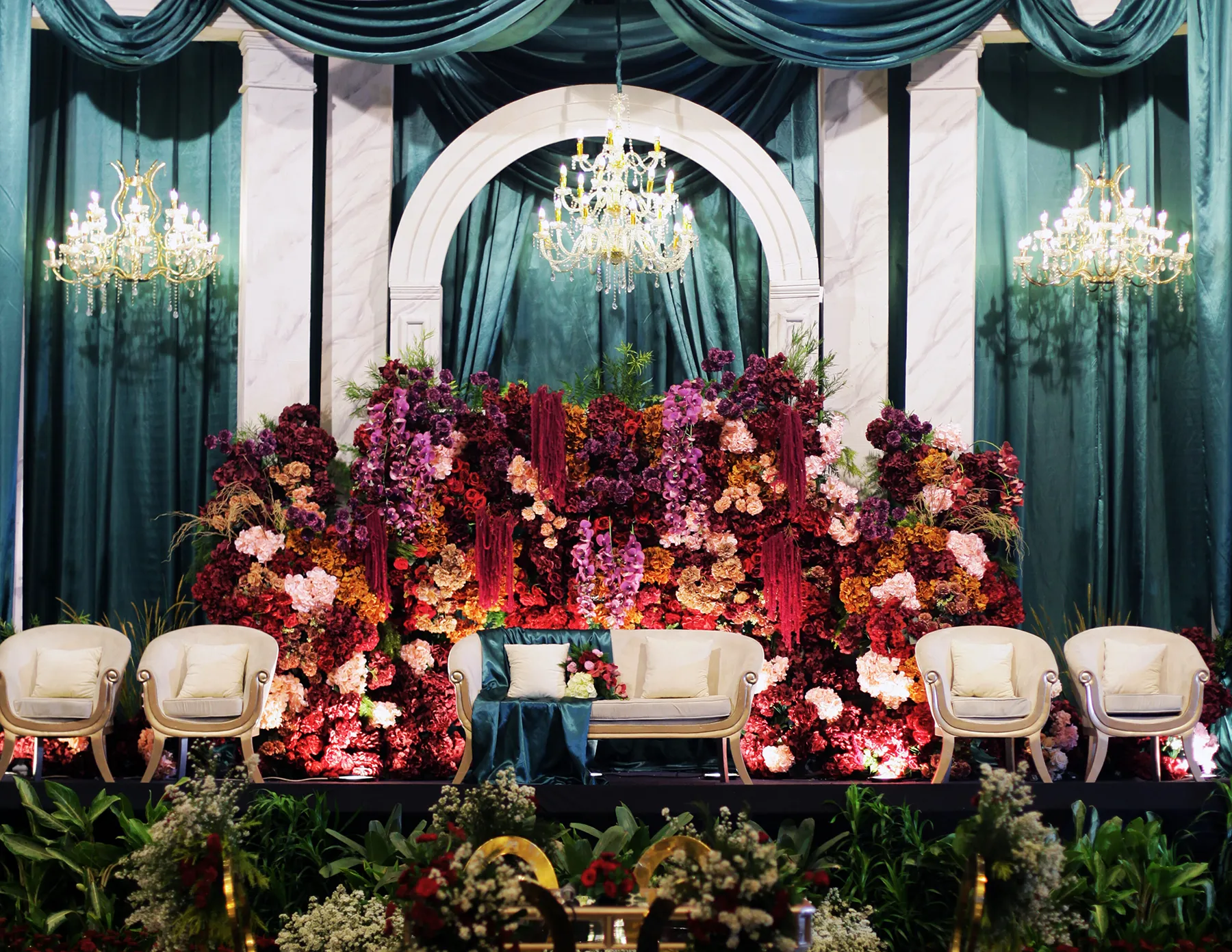 An opulent wedding reception stage featuring an abundant floral wall backdrop of burgundy, deep purple, pink, and amber blooms, framed by a white marble arch and dramatic teal satin drapes, with cream upholstered chairs and crystal chandeliers.