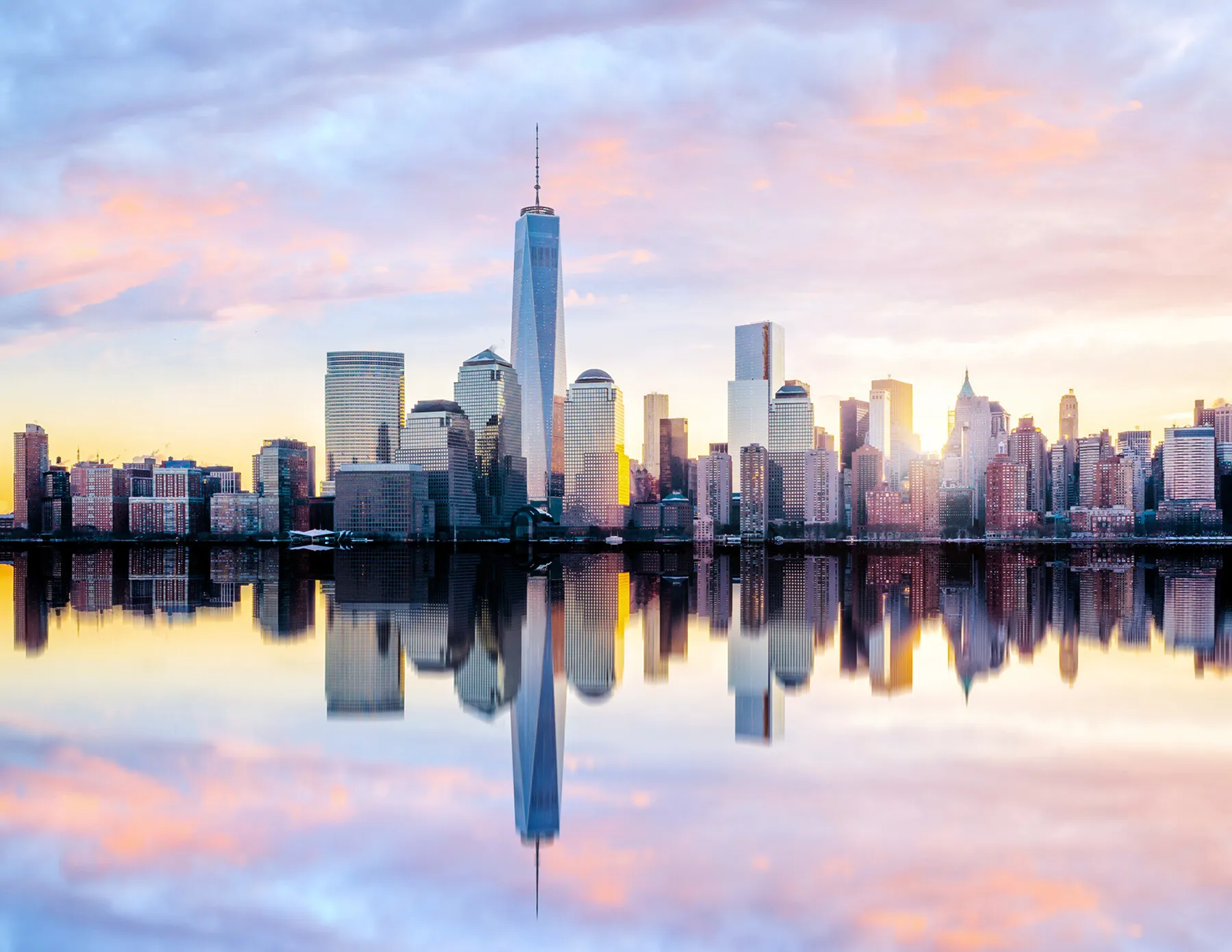 lower-manhattan-skyline-reflected-in-hudson-river-at-sunset-hmg-plus. The Lower Manhattan skyline dominated by One World Trade Center is perfectly reflected in the calm waters of the Hudson River beneath a pastel pink and purple sunset sky.