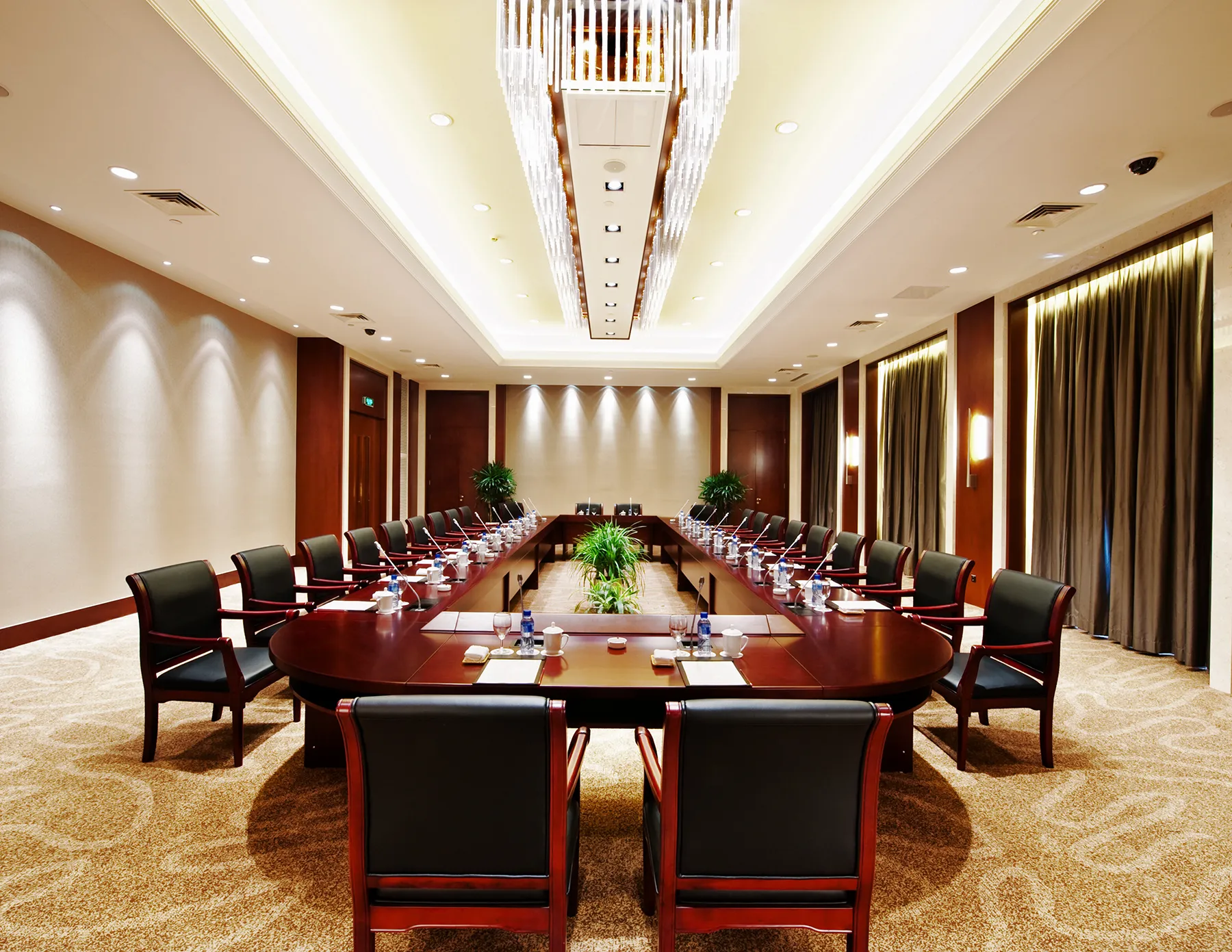 A large, elegantly appointed hotel conference room with a U-shaped mahogany table, black leather chairs, individual microphones, and a rectangular crystal chandelier overhead.