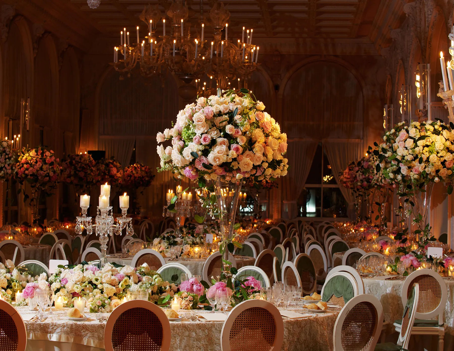 A grand ballroom wedding reception featuring towering arrangements of peach, pink, and cream roses on candlelit tables, beneath an ornate crystal chandelier.