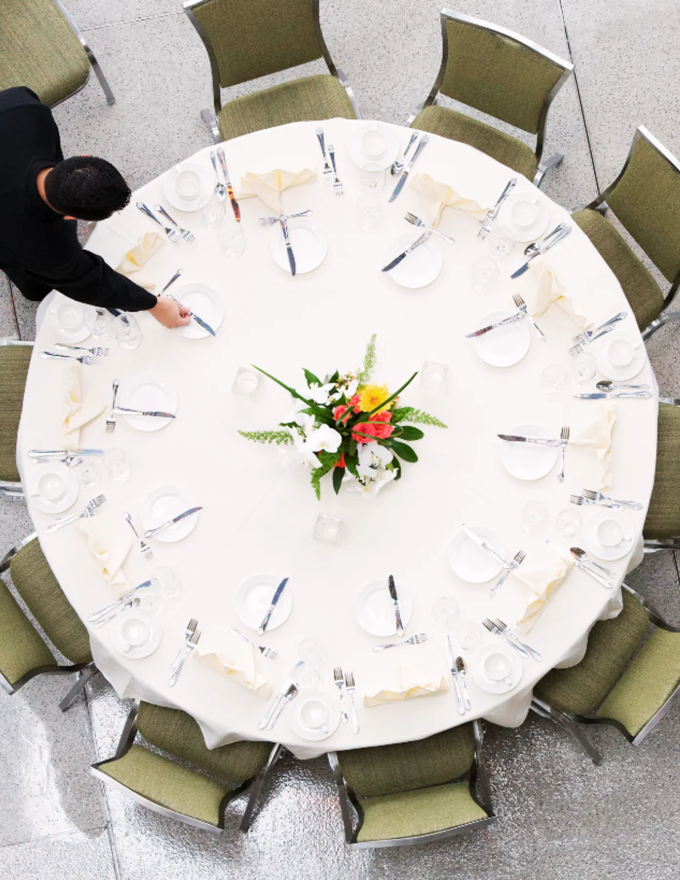 overhead-view-banquet-table-being-set-hmg-plus. Overhead view of a formally set round banquet table with a staff member placing settings, surrounded by green upholstered chairs.