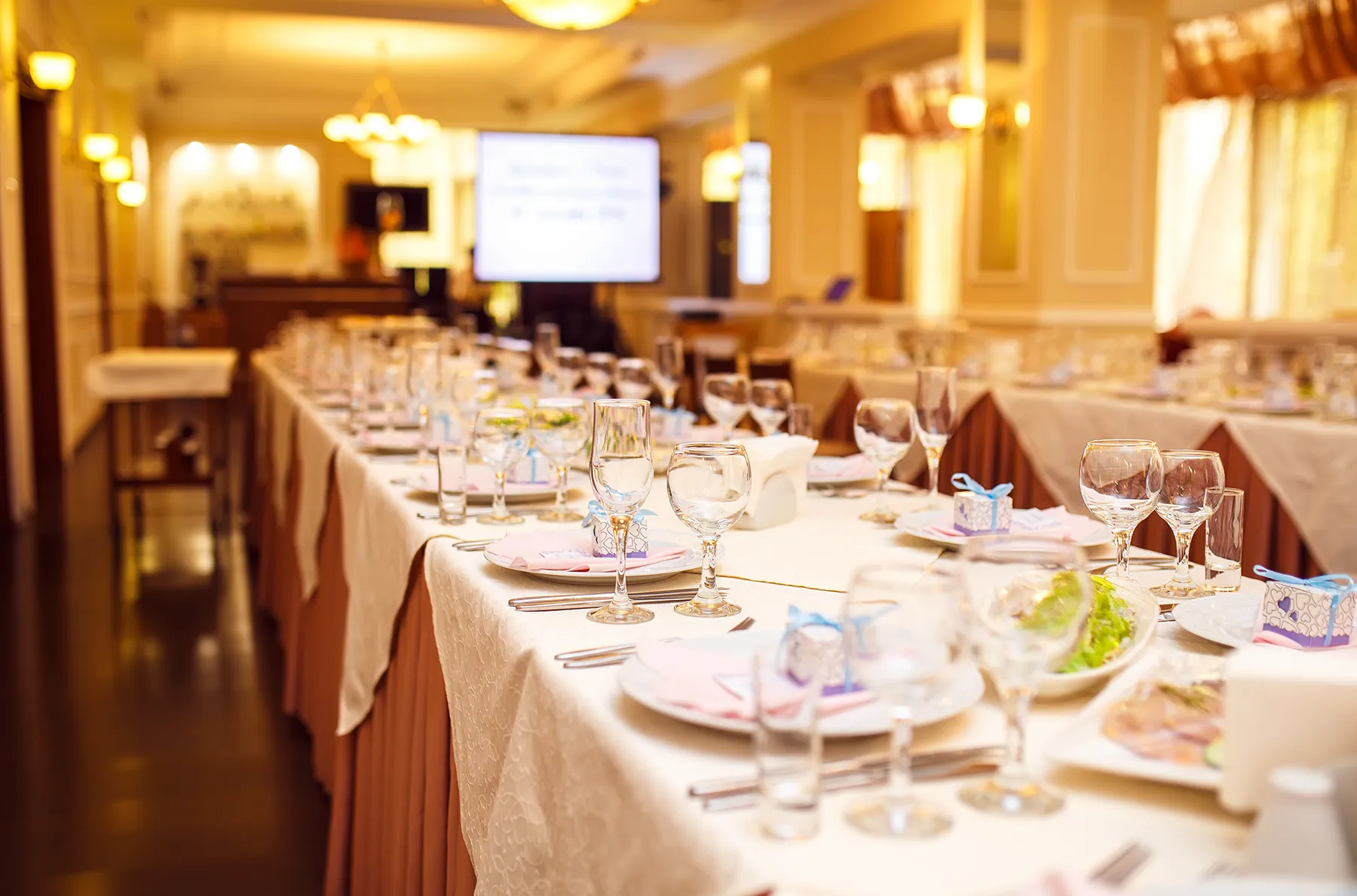 Elegantly set long banquet table with white linens, wine glasses, pink napkins, and party favors in a warmly lit event hall.