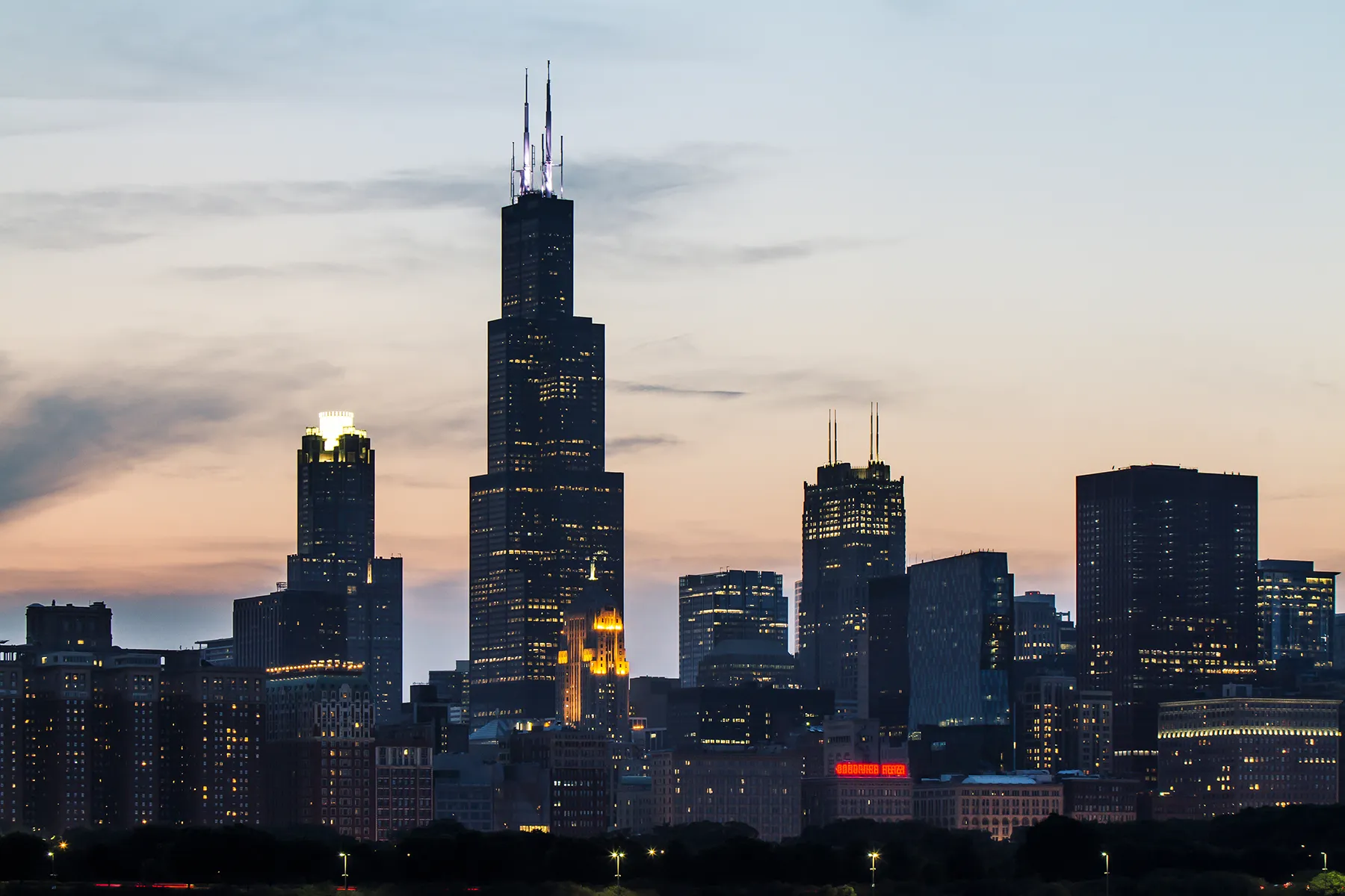 chicago-skyline-willis-tower-at-dusk-hmg-plus. Chicago skyline at dusk with Willis Tower prominently lit against a gradient sunset sky.