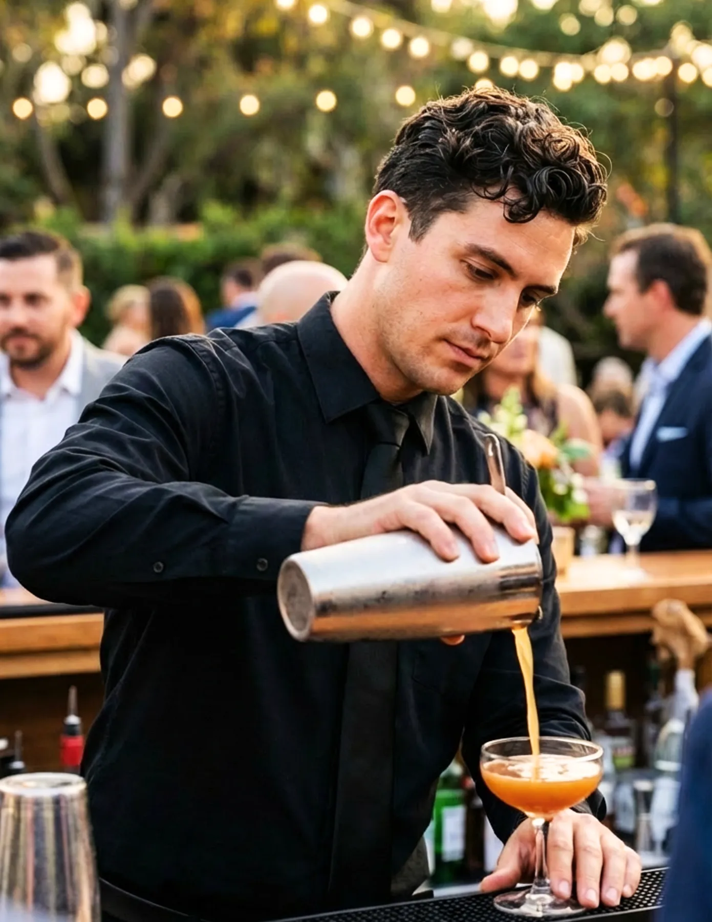 A focused male bartender in black pouring a cocktail from a shaker into a coupe glass at an outdoor evening event.