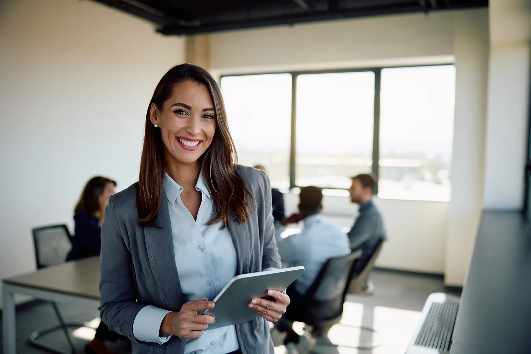 A smiling businesswoman in a gray blazer holding a tablet in a modern office conference room.