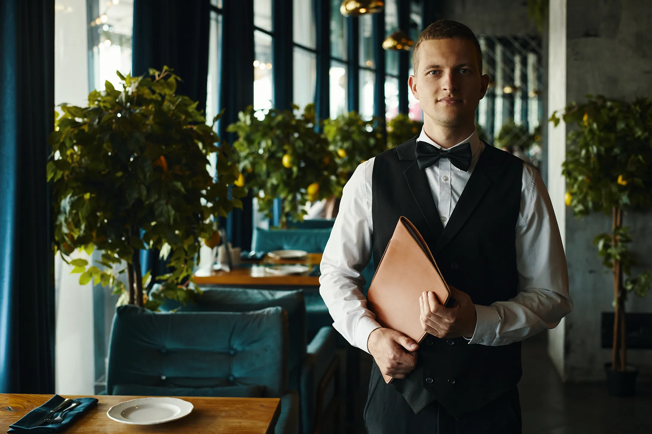 fine-dining-server-bow-tie-leather-menu-upscale-restaurant-hmg-plus. A male fine dining server in a black vest and bow tie holding a leather menu portfolio in an upscale restaurant.