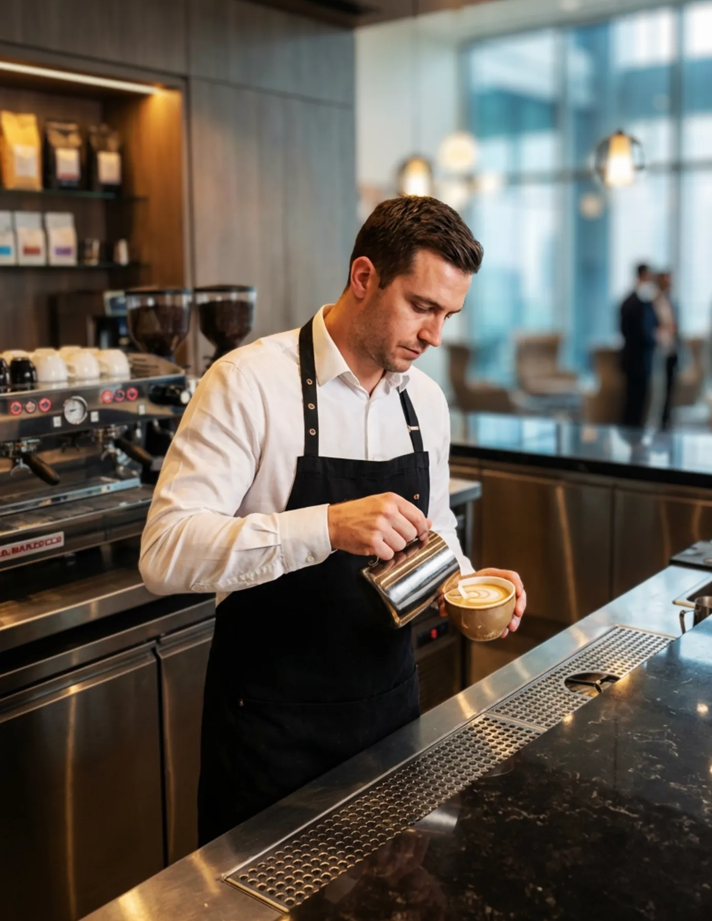 barista-pouring-latte-art-upscale-hotel-coffee-bar-hmg-plus. Barista wearing an apron pouring steamed milk to create latte art behind a modern café counter