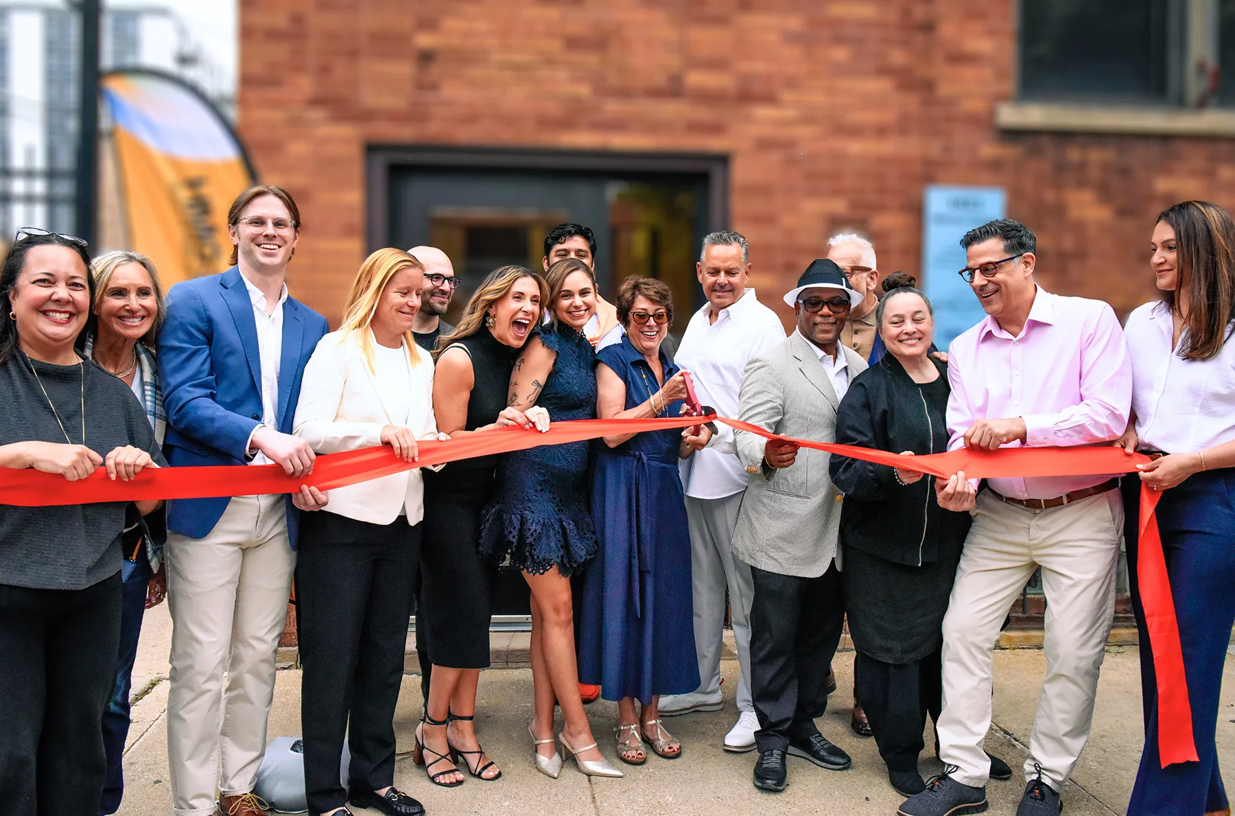 A large group of smiling people cutting a red ribbon together at a grand opening ceremony outside a brick building.