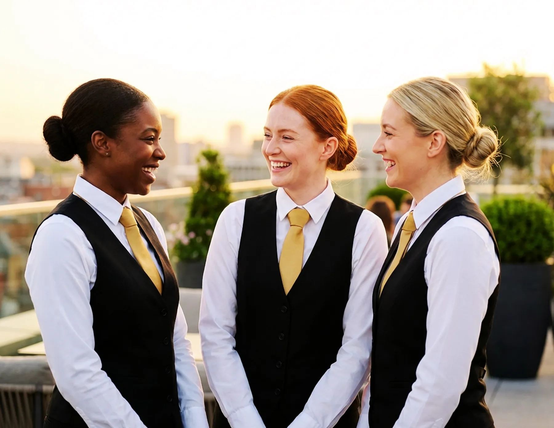 three-female-event-staff-gold-ties-rooftop-sunset-hmg-plus. Three female hospitality staff members in black vests and gold ties laughing together on a rooftop terrace at sunset.