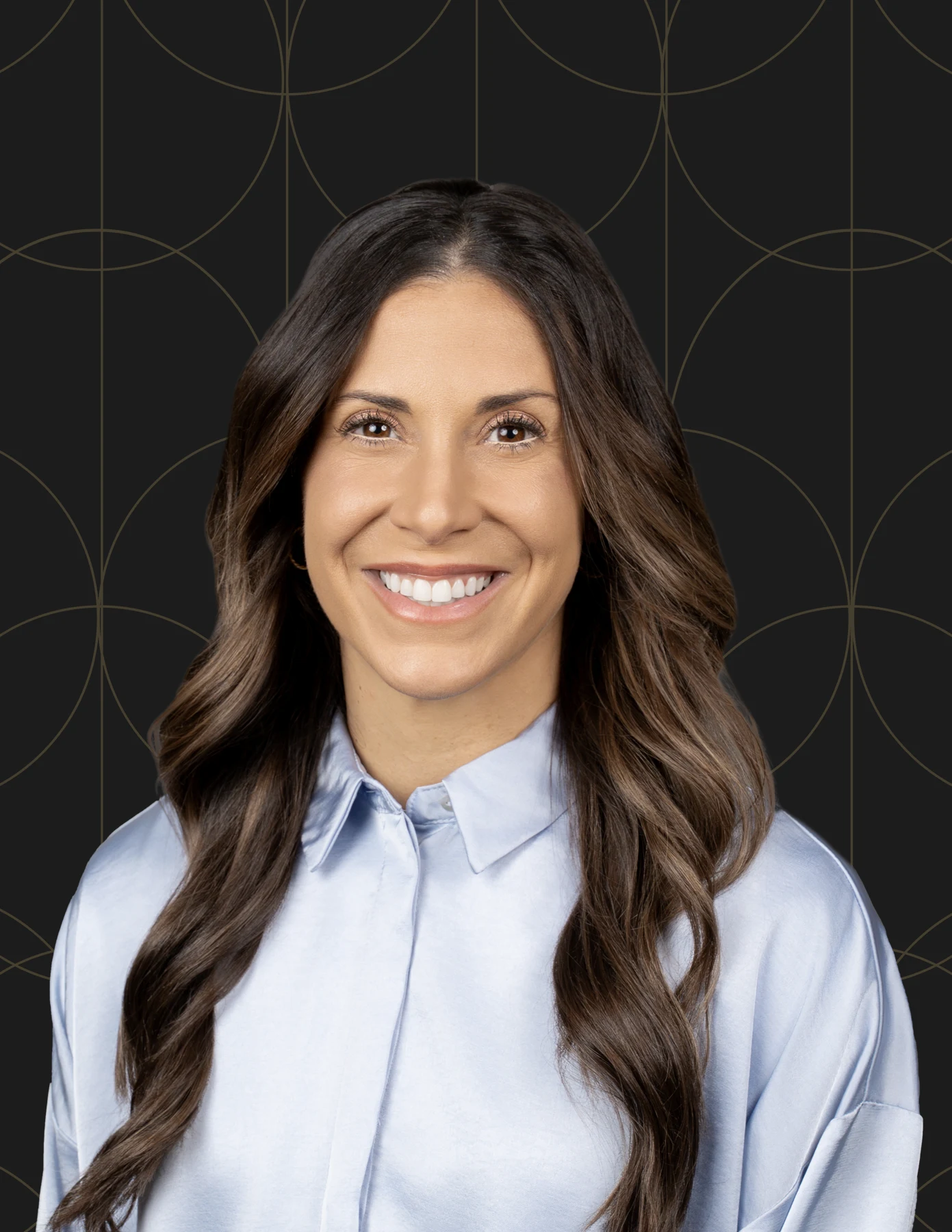 Professional headshot of a woman with long wavy dark brown hair wearing a light blue satin shirt.