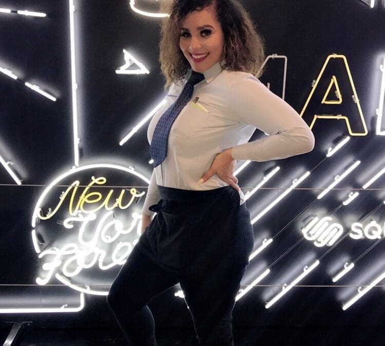 Female hospitality worker in a white dress shirt and tie posing confidently in front of a New York-themed neon light backdrop