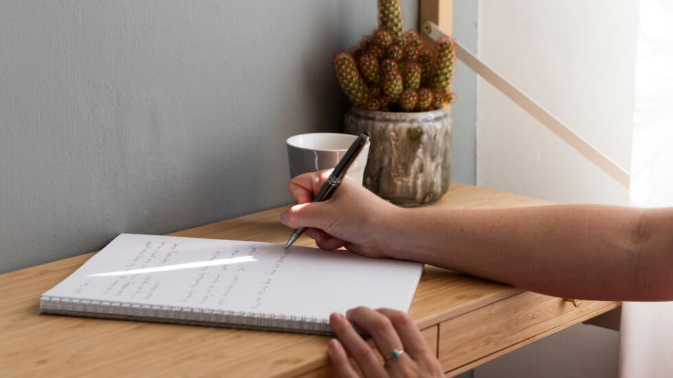A person's hand writing in an open notebook with a pen on a light wooden desk, with a small cactus plant and a cup visible in the background.