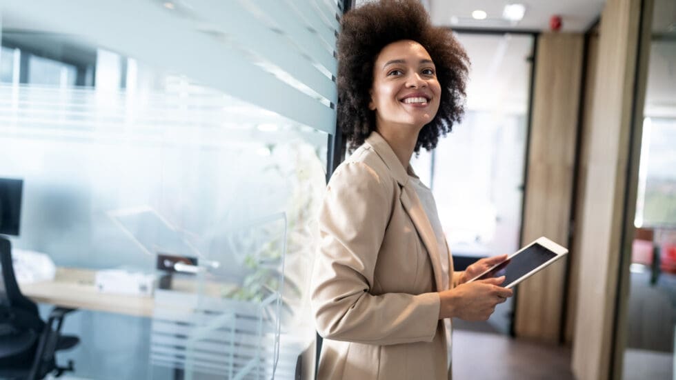 A confident young businesswoman with natural curly hair smiles over her shoulder while holding a tablet in a bright, modern office corridor lined with glass partitions.