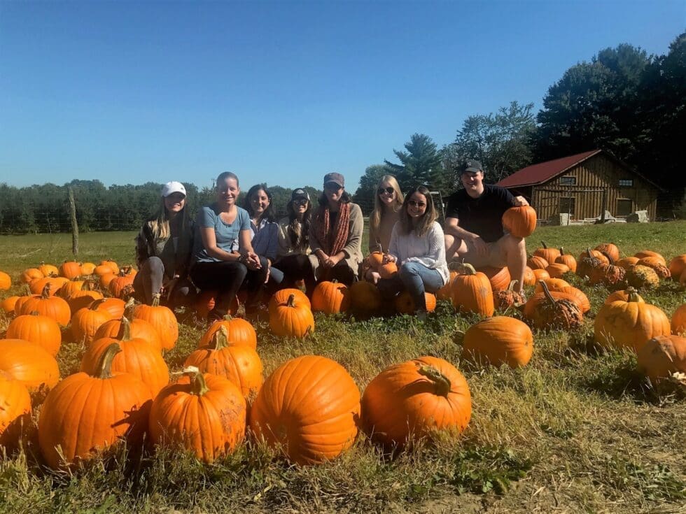 A group of eight smiling people pose among rows of bright orange pumpkins in a sunny autumn pumpkin patch, with a rustic barn and tree line visible in the background under a clear blue sky.