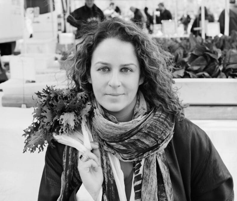 A black and white portrait of a curly-haired woman wearing a scarf and jacket, holding a bunch of fresh leafy greens at what appears to be an outdoor farmers market, with produce stalls and people visible in the background.
