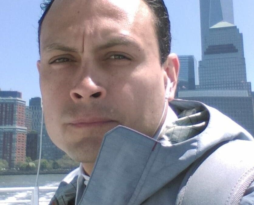 Man in a light gray suit squinting in bright sunlight on a boat with a New York City skyline in the background