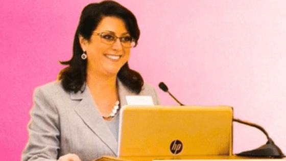 Smiling woman with glasses speaking at a podium with an HP laptop at a professional event against a pink background