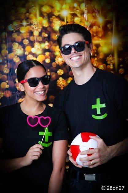 Two smiling people wearing matching black HMG+ t-shirts and sunglasses pose in front of a glittering gold bokeh photo booth backdrop, one holding a pink heart-shaped prop and the other holding a red and white ball.
