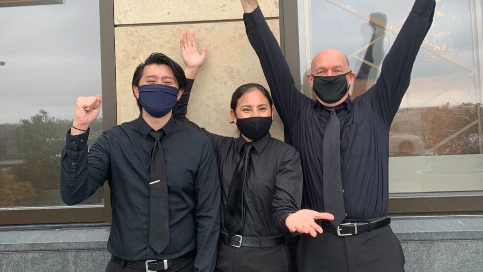 Three hospitality workers in black uniforms and face masks cheering and raising their arms outdoors