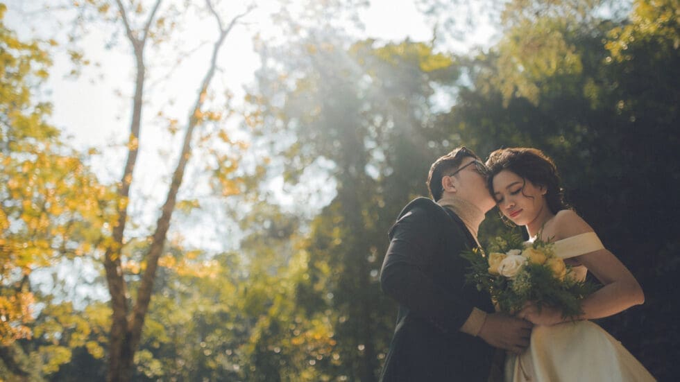 Groom kissing bride on the cheek in a sunlit forest setting, bride holding a bouquet of white and yellow flowers