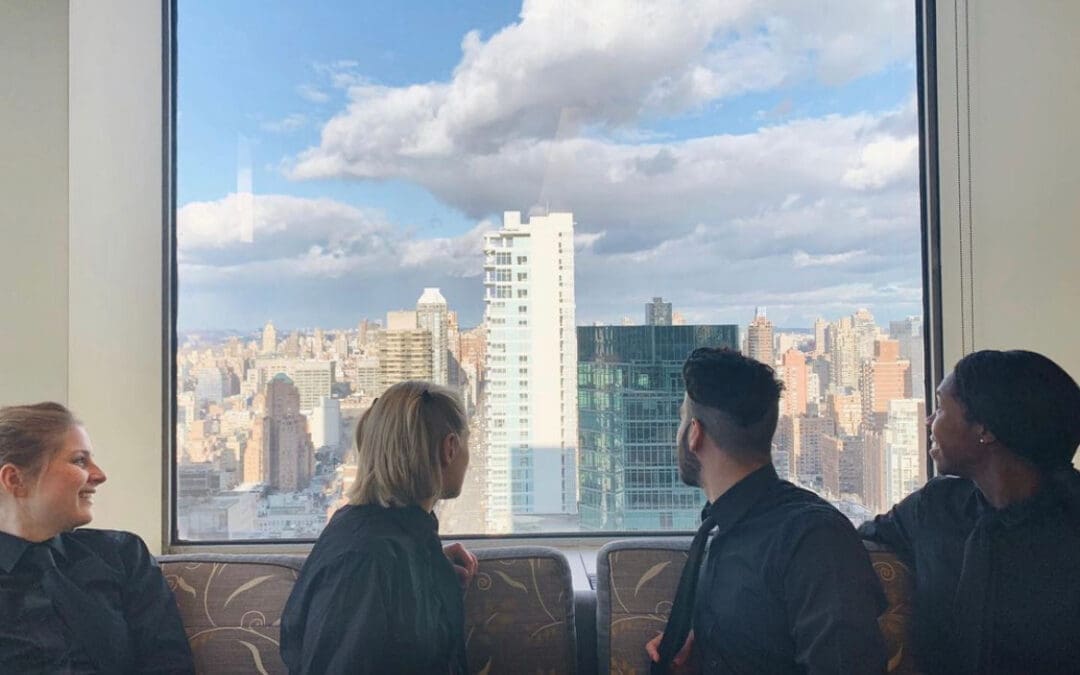 Four hospitality staff members in black uniforms relaxing and chatting near a large window with a panoramic New York City skyline view