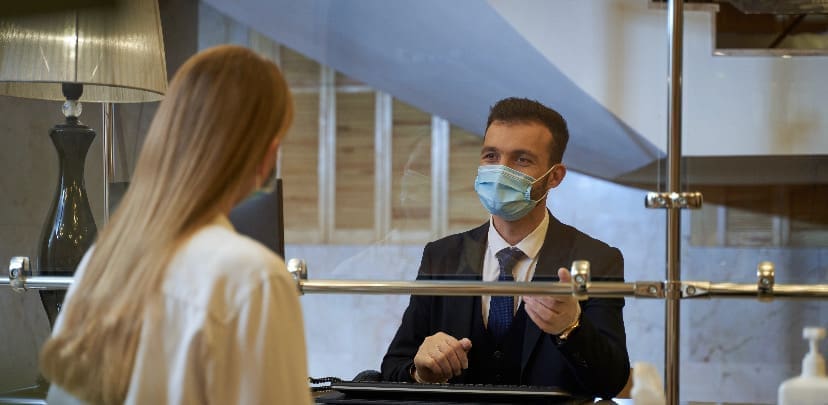 A masked male hotel receptionist in a dark suit assists a female guest at a front desk fitted with a protective screen, in a softly lit hotel lobby.
