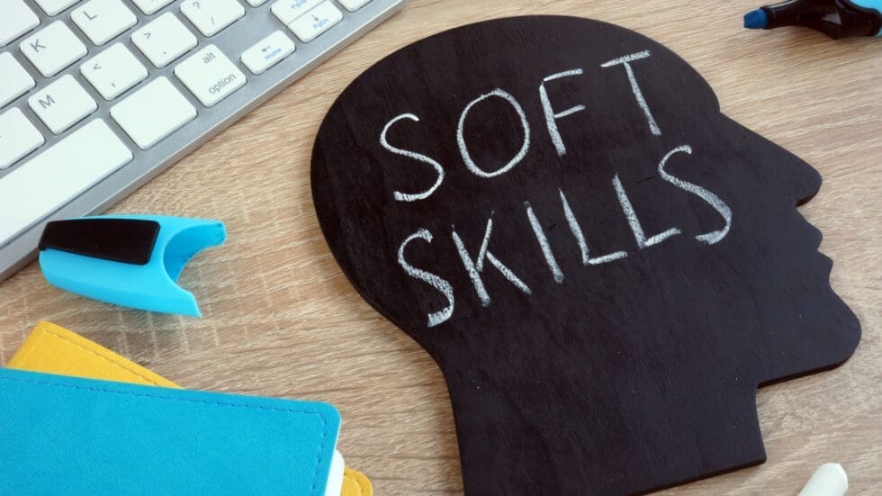 A black chalkboard cutout in the shape of a human head with the words "Soft Skills" written on it in white chalk, placed on a wooden desk alongside a keyboard, eraser, and colourful sticky notes.