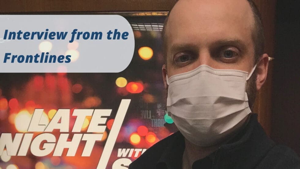 A masked man stands in front of a illuminated "Late Night" television show backdrop, with the text "Interview from the Frontlines" overlaid in the upper left corner of the image.