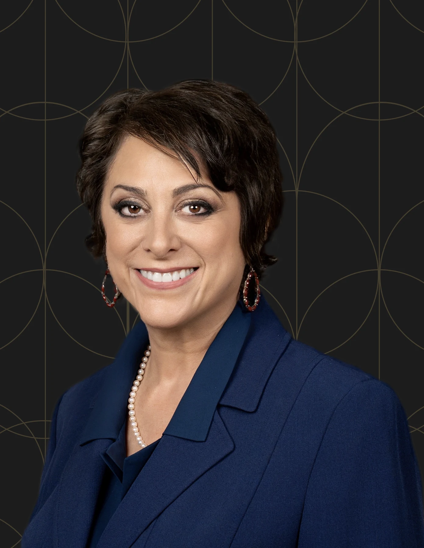 Professional headshot of a woman with short dark hair wearing a navy blazer, pearl necklace, and red hoop earrings.