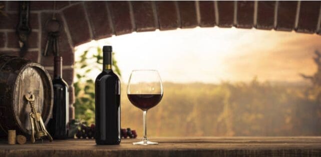A bottle and glass of red wine on a rustic stone ledge inside a brick archway, with a wooden barrel, corkscrew, and wine corks nearby, overlooking a sun-drenched vineyard in the background.