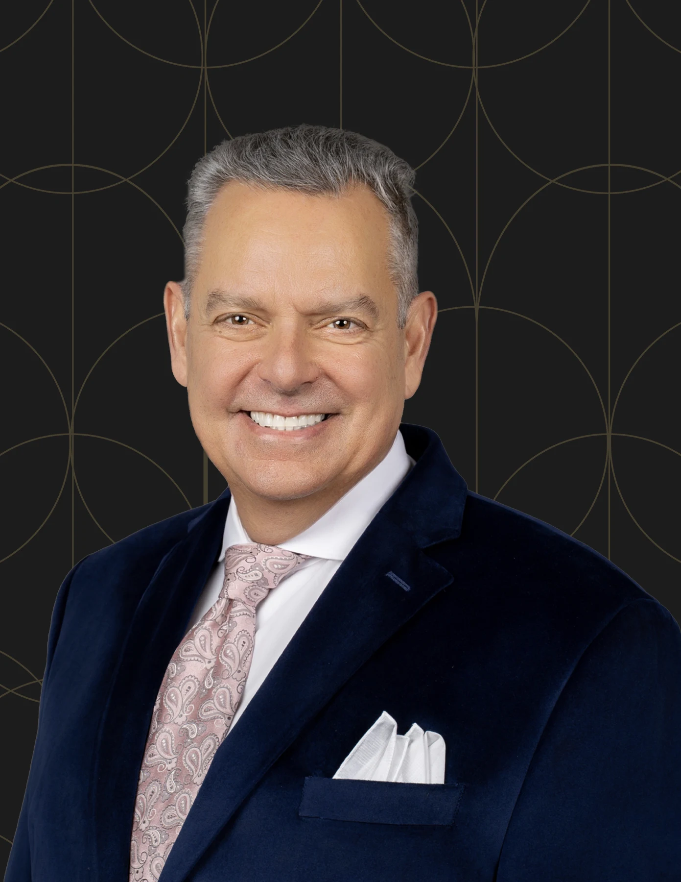 Professional headshot of a man with silver hair wearing a navy velvet blazer, pink paisley tie, and white pocket square.