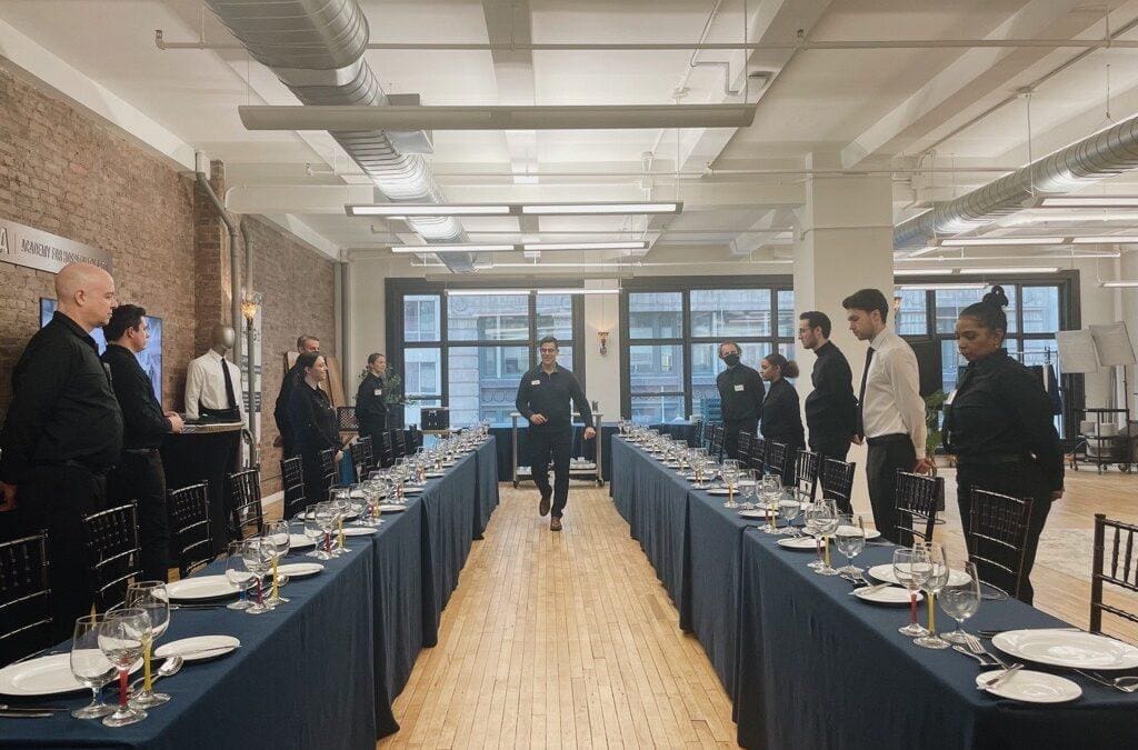 Event staff standing at attention along two formally set banquet tables in an industrial-style venue before a dinner service.