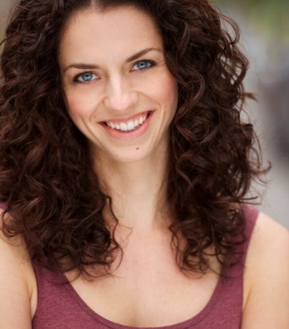 A professional headshot of a smiling woman with curly brown hair and blue eyes wearing a maroon top against a soft blurred background.