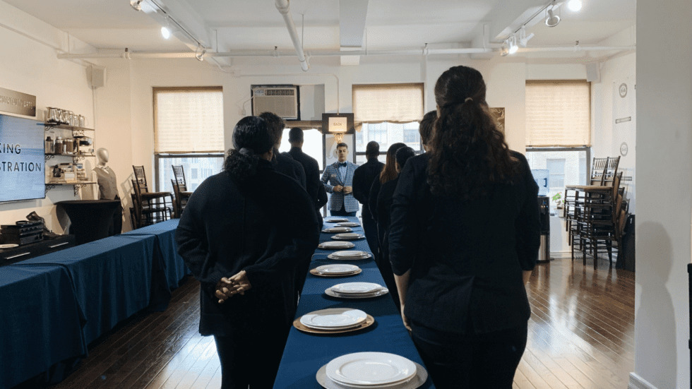A group of hospitality staff members stand around a long banquet table set with white plates and a blue tablecloth during what appears to be a training session in a bright event space.