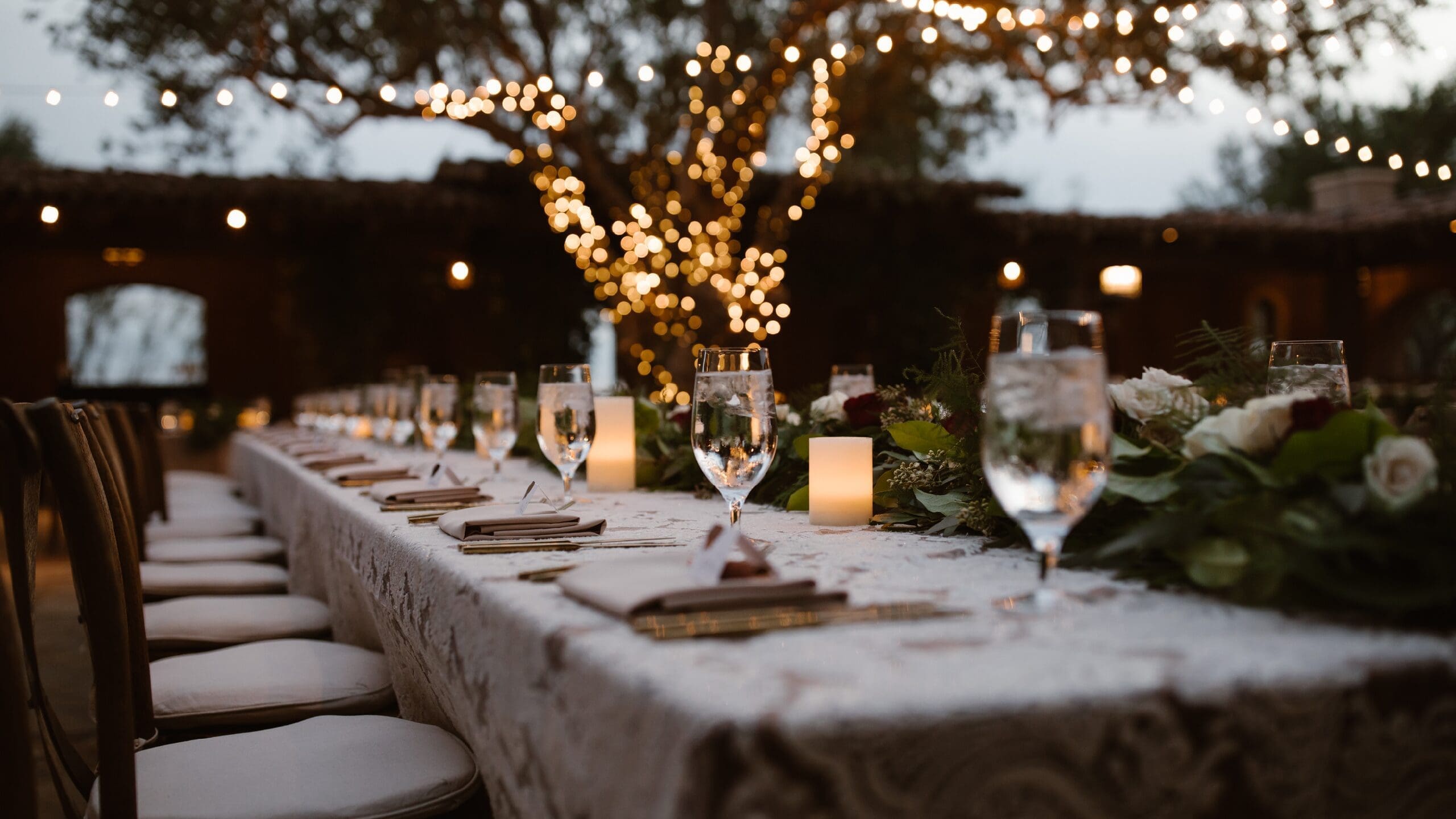 A long elegantly set outdoor banquet table with crystal glassware, candles, and floral greenery centrepieces, illuminated by warm fairy lights in the trees at dusk.
