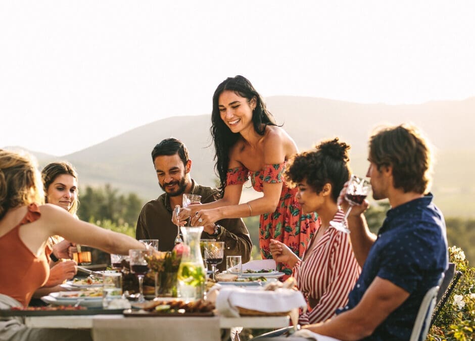 Group of friends dining and toasting at an outdoor table with a mountain landscape at golden hour