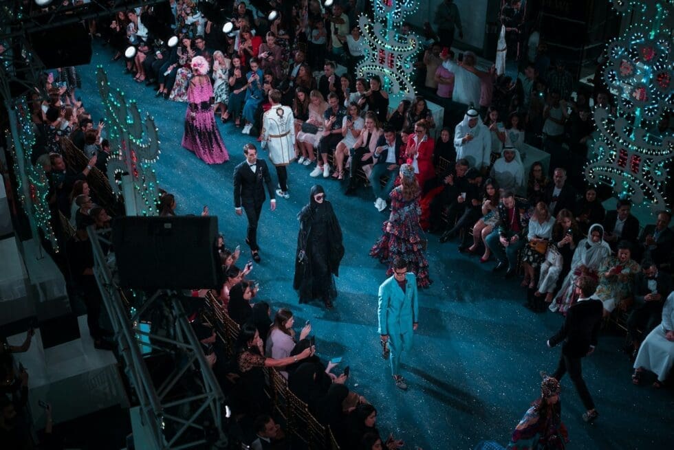 Models walking a glamorous fashion show runway surrounded by an engaged audience at a large-scale event