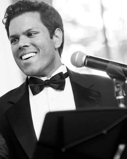 A black and white portrait of a smiling young man in a tuxedo and bow tie standing at a microphone and music stand.