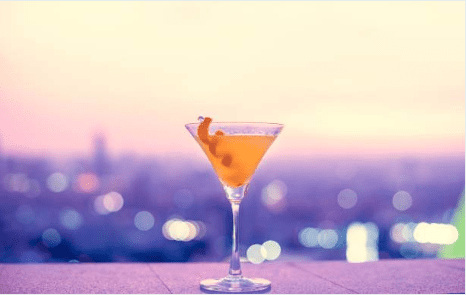 A golden cocktail in a martini glass garnished with a citrus twist, set against a soft, bokeh-lit city skyline at dusk in hues of pink and purple.
