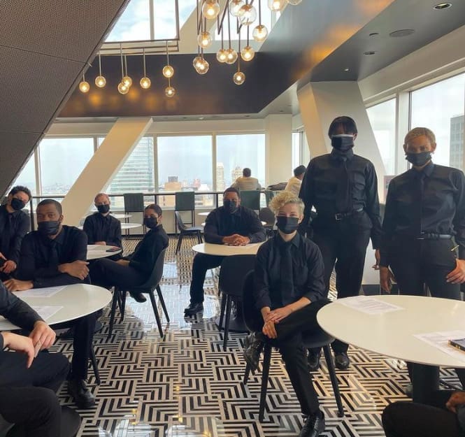 Group of masked hospitality staff in black uniforms seated and standing for a pre-event briefing in a modern high-rise venue