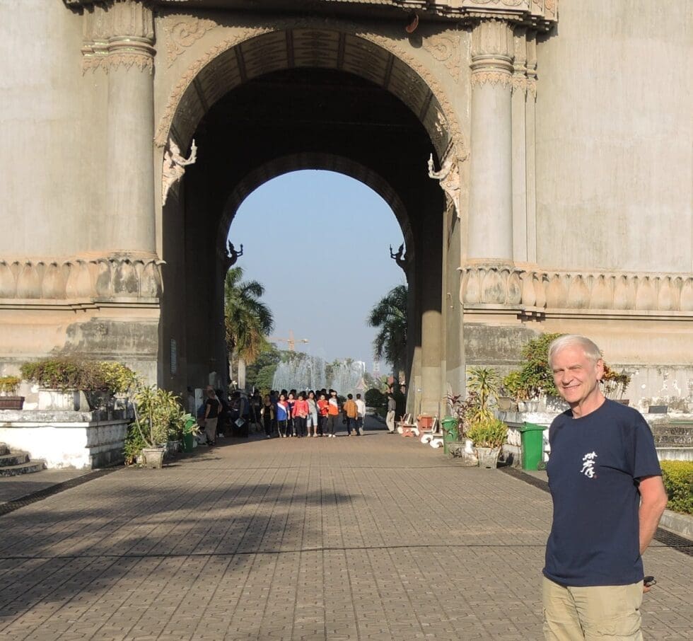 A smiling man (Terry Hanson) in a navy t-shirt and khaki pants stands in front of a large ornate stone arch monument, likely Patuxai in Vientiane, Laos, with a paved walkway, tropical plants, a fountain, and a crowd of visitors visible through the archway under a clear blue sky.