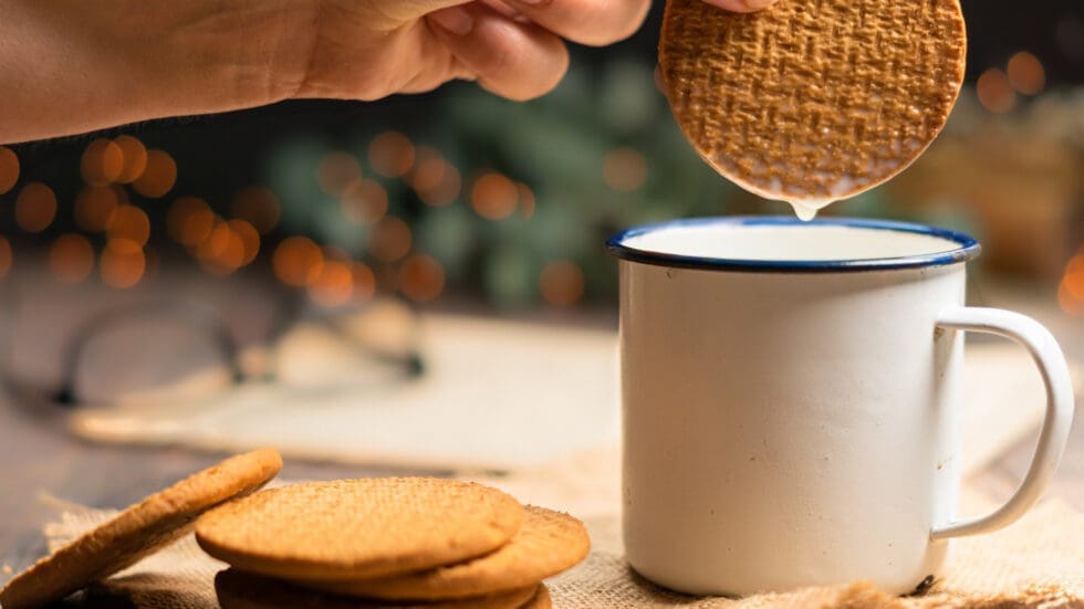 Hand dipping a round stroopwafel cookie into a white enamel mug of milk, with more cookies on burlap beside it and bokeh lights in the background