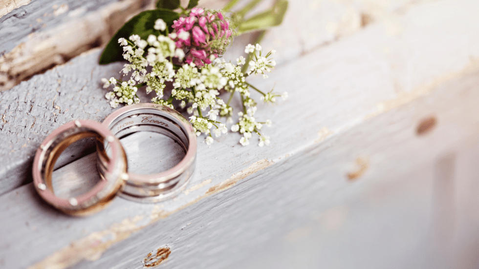 Two engraved wedding bands resting together on a weathered white wooden surface beside a small bouquet of white and pink wildflowers.