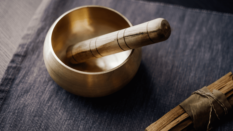 A golden Tibetan singing bowl with a wooden mallet resting inside it, placed beside a wrapped smudge stick on a dark textured linen surface.