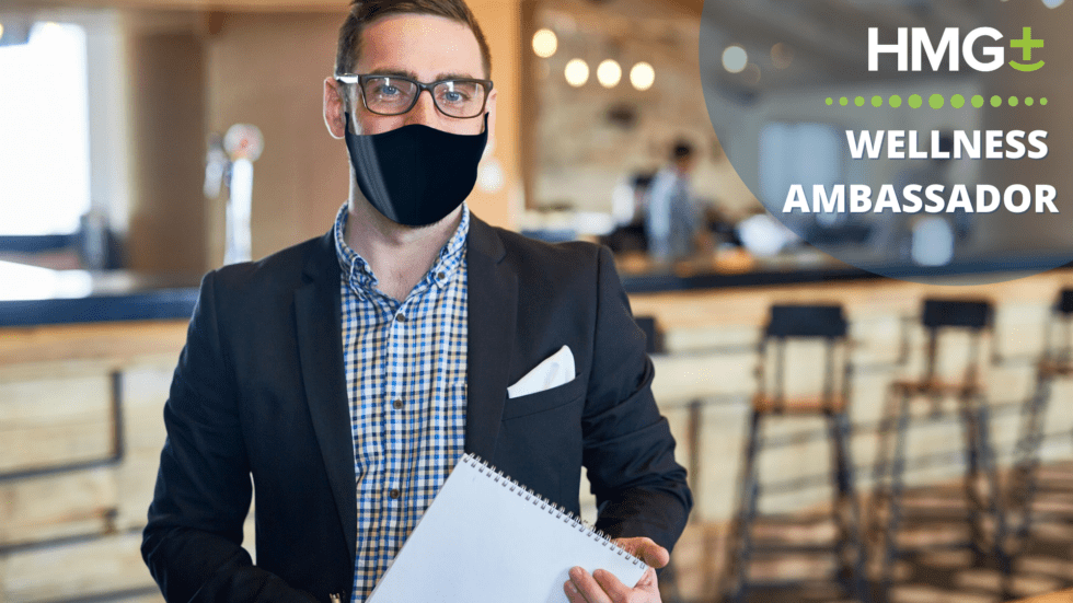 A masked male professional in a dark blazer holds a notebook and stands confidently in a hospitality venue, beside an HMG+ Wellness Ambassador logo overlay.