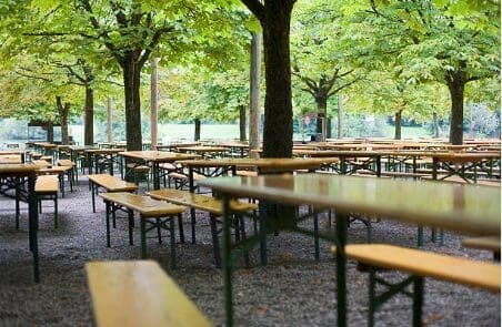 Rows of empty wooden tables and benches in an outdoor beer garden, shaded by a canopy of lush green trees on a gravel ground.